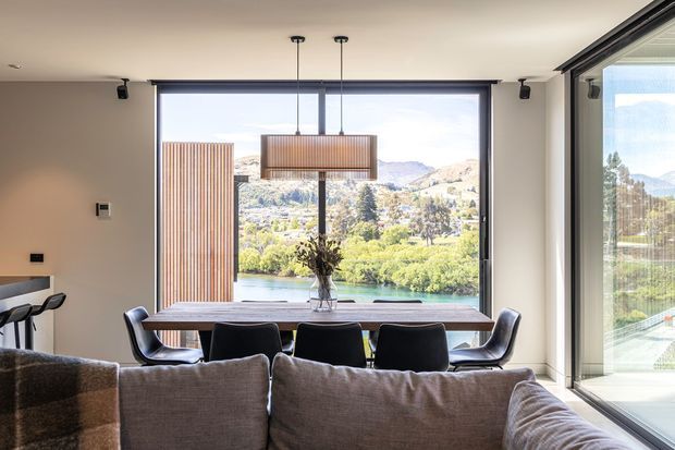A bright contemporary open-plan living and dining area viewed from behind a large sectional sofa. The dining zone features a long timber table, dark upholstered chairs, and a linear pendant light, while expansive black-framed glazing provides a panoramic view of a lake, landscaped surroundings, and hills. Neutral finishes and streamlined built-in elements create a refined, airy interior.