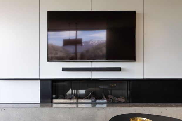 A minimalist contemporary living room feature wall with a large wall-mounted television above a linear built-in fireplace. The wall is clad in pale smooth panels with fine vertical joints, contrasted by a glossy black fireplace surround and dark hearth. A concrete-look floor and low black coffee table are visible in the foreground. The television reflects a mountainous landscape and pendant light, but the main subject is the clean, integrated media and fireplace wall.