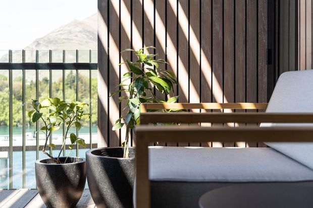 A sunlit balcony or terrace with dark timber slat screening, a black metal-and-glass balustrade, and potted leafy plants. A timber-framed outdoor lounge chair with light upholstery occupies the foreground, with a hazy mountainous landscape visible beyond the railing.