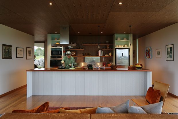 Warm, open-plan kitchen viewed from the living area, with a long island featuring a vertically ribbed light facade and timber benchtop. The kitchen has extensive dark timber cabinetry and open shelving, integrated appliances, a stainless-steel rangehood, pendant lights, and a timber-lined ceiling. Timber flooring, framed artwork, and a partially visible sofa and armchair create a relaxed contemporary living environment.