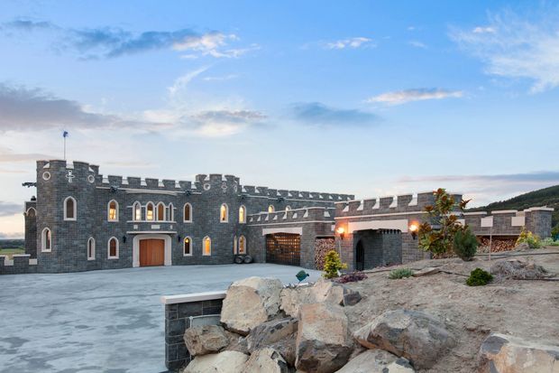 Exterior view of a large castle-inspired stone building set on a landscaped rocky site. The dark grey masonry facade features crenellated parapets, corner turrets, arched and narrow windows, timber entry doors, and warm exterior lighting. A broad paved forecourt and low stone retaining walls occupy the foreground, with rolling terrain visible beyond.