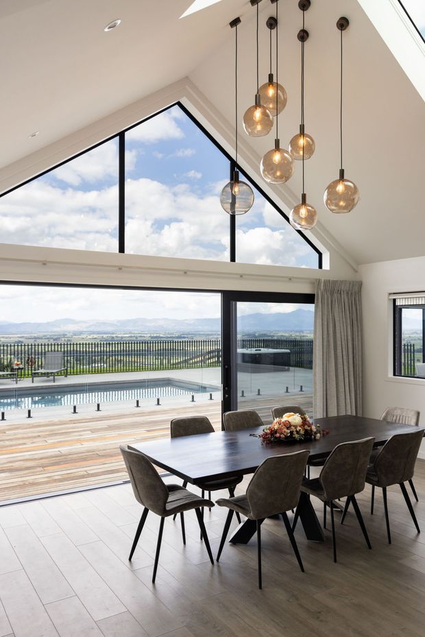Bright, contemporary dining area with a large dark rectangular dining table and upholstered chairs, positioned beside expansive black-framed glazing and sliding doors. A dramatic pitched ceiling with a triangular clerestory window brings in abundant daylight, while multiple suspended glass globe pendant lights hang at varied heights. Through the glazing is a timber deck, swimming pool, landscaped grounds and distant mountain scenery.
