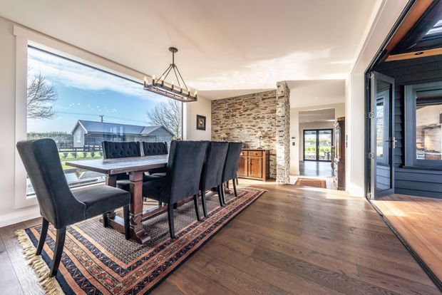 A bright dining area with a long dark wood dining table and upholstered black dining chairs arranged on a patterned rug. The room features wide floor-to-ceiling glazing, warm timber flooring, a rustic exposed-stone feature wall with built-in timber cabinetry, and a large glazed opening to an exterior or covered outdoor space. A linear pendant light hangs above the table, and an adjoining hallway is visible beyond.