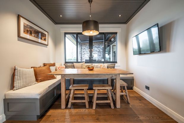 A compact, warm dining room with a built-in upholstered bench, rustic timber dining table and benches, and a wall-mounted television. The room has dark timber flooring, a dark painted timber-panel ceiling, pale walls, a black pendant light, framed artwork, and large windows looking toward a stone-clad exterior area.