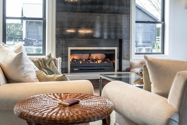 Bright contemporary living room centered on a modern built-in fireplace with a dark textured feature wall. Large windows flank the fireplace, while neutral upholstered sofas and armchairs surround a glass coffee table and woven round ottoman.
