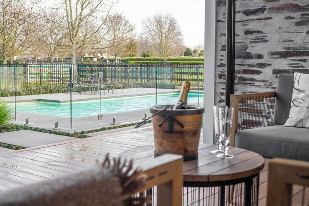 A contemporary outdoor entertaining area viewed from inside, with a timber deck, round timber dining table, woven and upholstered seating, and a stone-clad wall. Full-height glazing opens to a fenced swimming pool and landscaped grounds beyond.