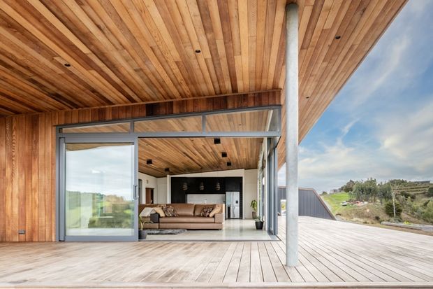 Modern timber-clad home with a broad covered deck and open-plan living area visible through large sliding glass doors. The ceiling, walls, and deck are lined in warm timber, with exposed structural posts, dark-framed glazing, and a partially visible kitchen and lounge. The building sits in a semi-rural landscaped setting.