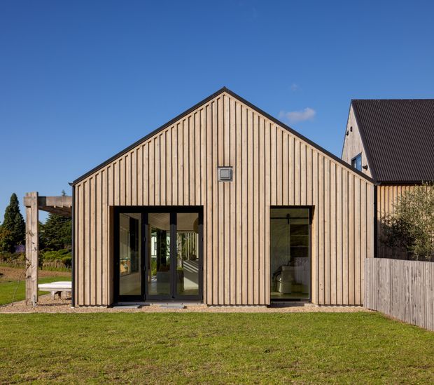 Exterior view of a simple gabled single-storey building clad in vertical light timber boards, with dark-framed glazed doors and windows. The structure sits beside a lawn and timber fencing, with a larger pitched-roof building visible adjacent to it. Clear blue sky and restrained detailing give the facade a contemporary barn-like character.