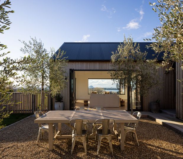 A landscaped outdoor dining courtyard with a large timber dining table and metal chairs, opening toward a contemporary pavilion-style house. The building has dark corrugated metal roofing, vertical timber cladding, extensive glazed openings, and a warm neutral interior visible through the open doors. Mountain scenery is visible through the rear glazing, with mature olive trees and gravel landscaping surrounding the entertaining area.