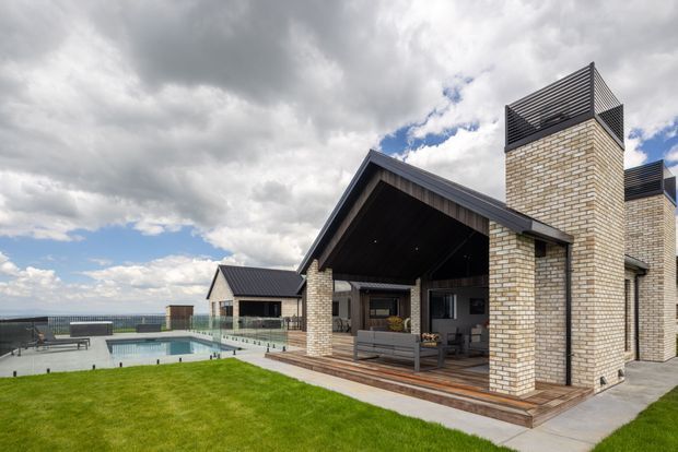 Exterior view of a contemporary single-storey home with a prominent gabled pavilion, dark metal roof, pale brick chimneys, timber-lined covered outdoor living area, glass pool fencing, and a swimming pool beside a manicured lawn. The property appears elevated with a broad coastal or rural horizon beyond, under a dramatic cloudy sky.