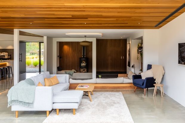 A bright contemporary open-plan living room with a large pale sectional sofa, blue accent armchair, timber coffee table, and light rug on a polished concrete floor. The room features a warm timber-lined ceiling, dark timber built-in cabinetry and fireplace surround, white walls, a partially visible kitchen, and large glazed openings to the garden.