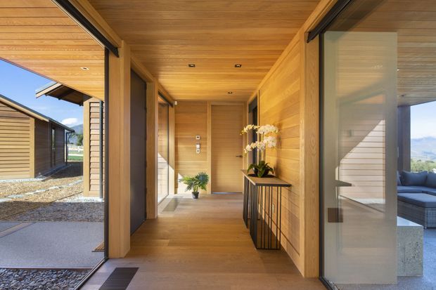 A warm, timber-lined residential entry hall with full-height glazed sliding doors opening to an outdoor courtyard and mountain views. The corridor features a timber ceiling and walls, recessed downlights, timber flooring, a narrow black-framed console, and potted plants. Adjacent rooms and a sheltered outdoor seating area are visible through the glazing.