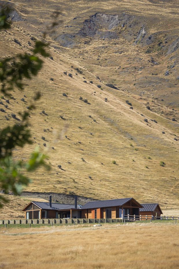 A low, single-storey rural house sits at the base of a steep, dry grass-covered mountainous hillside. The home has dark corrugated pitched roofs, warm timber-clad walls, extensive glazing, covered verandas, and a fenced foreground pasture. The scene has a remote alpine or high-country rural character.
