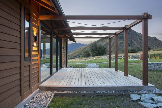 Exterior view of a timber-clad building opening onto a broad timber deck and pergola structure. The deck has slim wooden posts and horizontal beams with tension cables, while full-height glazed doors connect the interior to the outdoor space. A landscaped rural setting with gravel, low stone edging, fencing, and mountainous terrain is visible beyond.