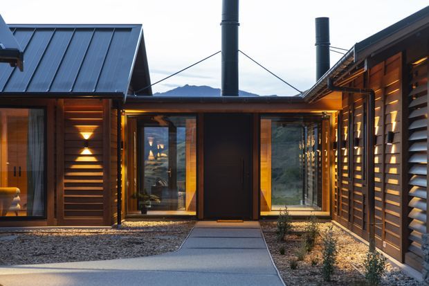 A compact contemporary single-storey residential entry courtyard framed by dark timber-clad wings. The central entrance has a solid dark door, warm timber-framed glazing, and a concrete path leading through gravel landscaping. Slatted timber screening, exposed roof forms, and tall dark metal flues are prominent, with mountainous terrain visible beyond the building.