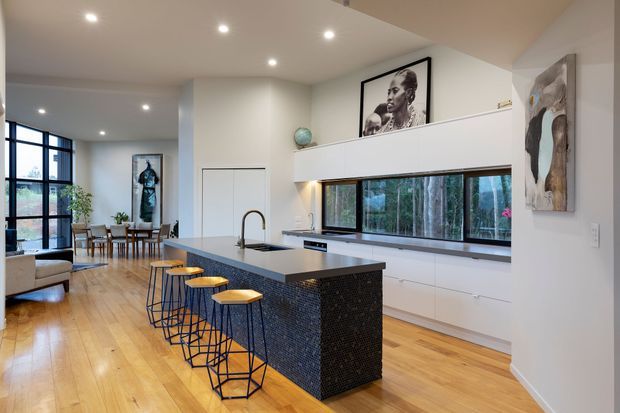 Bright open-plan contemporary kitchen with a large dark tiled island, integrated sink and seating for four, opening toward a dining and living area. Light timber flooring, white cabinetry, recessed lighting, expansive black-framed glazing, and a long horizontal window create a clean, airy interior. Artwork and a timber-lined outlook add visual interest.