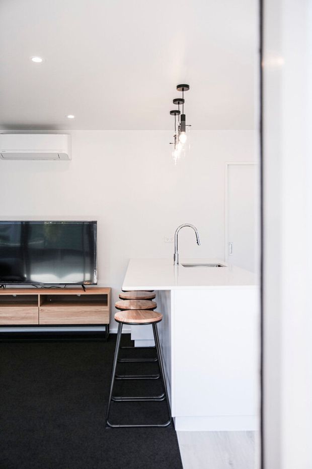 A compact open-plan living and kitchen area viewed from a doorway. The room has white walls and ceiling, a white island bench with an integrated sink and curved chrome tap, three timber-topped black stools, a timber TV console, wall-mounted television, recessed lighting, and a small cluster of pendant lights. Dark carpet contrasts with pale timber flooring visible near the kitchen.
