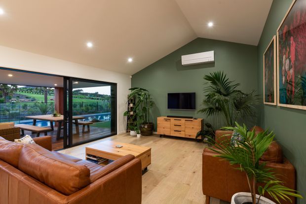 Warm contemporary living room with a cognac leather sectional sofa, timber coffee table and media console, green feature walls, indoor palms and framed artwork. Large sliding glass doors open to an outdoor terrace and landscaped view, while recessed lighting and a wall-mounted air conditioner complete the room.