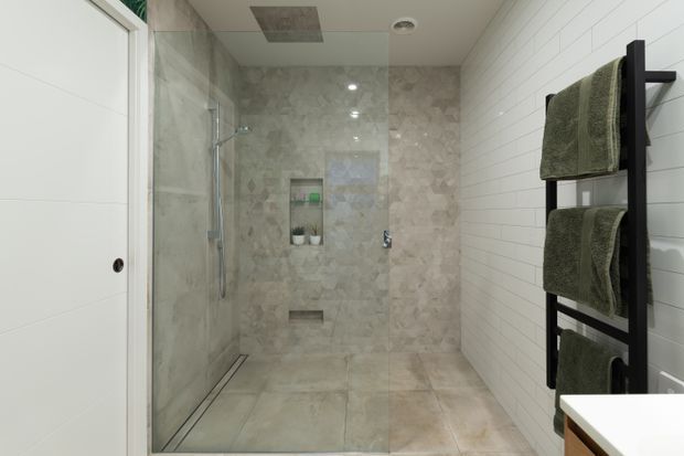 A contemporary walk-in shower bathroom with a frameless glass enclosure, large pale stone-look floor and shower wall tiles, and a white horizontal subway-tile wall. A black ladder-style heated towel rail with olive-green towels is mounted beside a white vanity.
