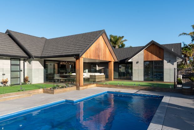 Exterior view of a contemporary single-storey home arranged around a swimming pool. The building has multiple gabled roof volumes, dark grey tiled roofing, light grey masonry walls, warm timber-clad gable ends, expansive glazing, and a paved pool terrace with landscaped lawn.
