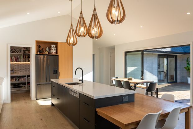 Bright contemporary open-plan kitchen and dining area with a large dark island, pale stone benchtop, timber cabinetry, integrated stainless-steel refrigerator, walk-in pantry, pendant lighting, and timber dining tables. Full-height glazing opens to an outdoor courtyard or patio.