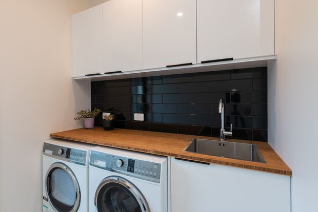 A compact, modern laundry room with white overhead cabinetry, a timber benchtop, glossy black subway-tile splashback, stainless-steel sink and tap, and two front-loading washing appliances beneath the counter. Small potted plants add a subtle decorative touch.