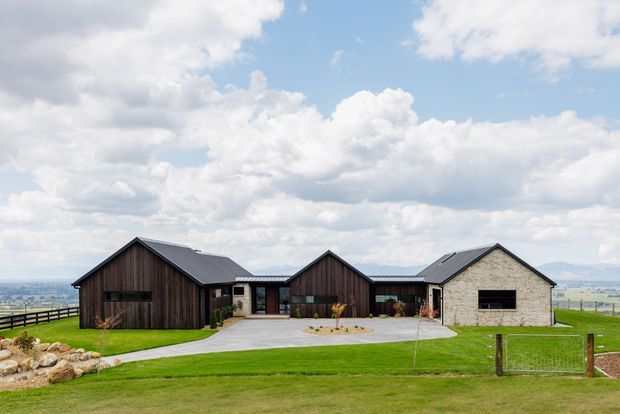 Single-storey contemporary rural residence viewed from the front, composed of several connected gabled volumes. The dark vertical timber-clad wings contrast with a pale masonry central/right wing and dark pitched metal roofs. A broad concrete driveway and entry path lead through manicured lawns, with fencing and an expansive open rural landscape beyond.