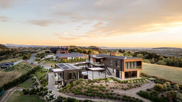 Aerial view of a contemporary multi-building residential property set on a landscaped, elevated rural-suburban site. The complex features dark rectangular volumes, extensive glazing, flat roofs, timber-toned facade accents, terraces, pathways, and landscaped gardens, with rolling terrain and distant development beyond.