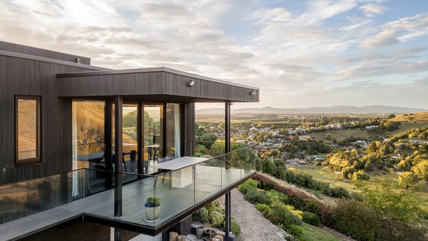 A modern elevated home overlooking a broad suburban and rural landscape. The dark vertical-clad facade has a flat roof, extensive floor-to-ceiling glazing, and a projecting glass-balustraded terrace or balcony with outdoor furniture. The view beyond includes rolling hills, gardens, and a settled valley under a partly cloudy sky.