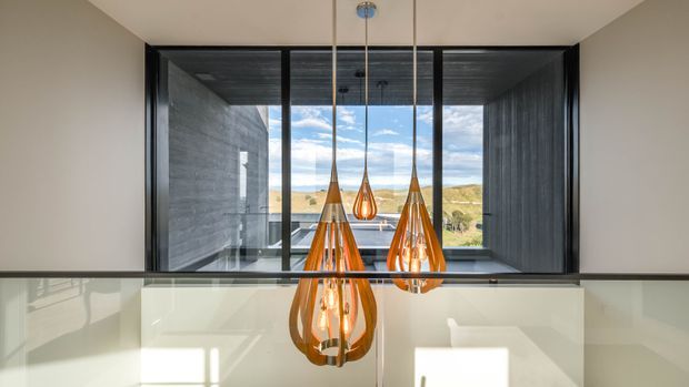 Interior view from an upper-level landing or hallway overlooking a stairwell, with a clear glass balustrade in the foreground. Three sculptural amber pendant lights hang in front of expansive black-framed glazing, revealing a broad rural landscape beyond. The space has a restrained, contemporary architectural character with dark timber or textured ceiling and wall surfaces, pale walls, and strong indoor-outdoor visual connection.