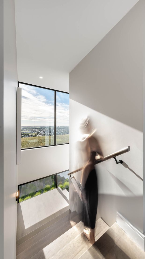 A bright, minimalist interior stairway with pale wood or stone-look treads, white walls, and a simple metal handrail. Large black-framed windows bring in strong natural light and overlook a suburban landscape. A blurred person is descending the stairs, emphasizing the vertical circulation space and dramatic sunlight and shadows.