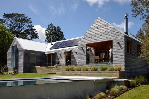 Exterior view of a contemporary single-storey home with several steep gabled volumes clad in weathered grey timber boards. Large recessed openings create covered outdoor living areas, while a broad deck overlooks a raised rectangular swimming pool. Solar panels are mounted on the roof, and a metal flue rises from one gable. The house sits within a landscaped suburban setting with mature trees and ornamental grasses.
