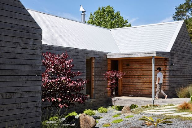 Exterior view of a contemporary single-storey dwelling with dark horizontal timber cladding, bright corrugated metal gabled roof forms, and a recessed timber-lined entry porch. The landscaped approach uses pale gravel, large stepping stones, boulders, grasses, and ornamental planting.