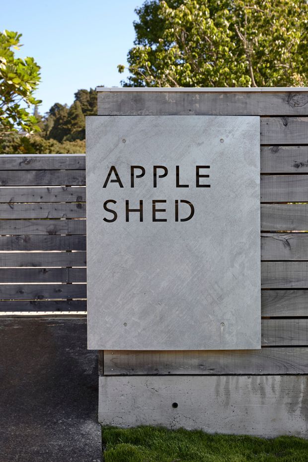 Exterior wayfinding sign reading “APPLE SHED” mounted on a weathered horizontal timber privacy fence above a raw concrete base. The scene is outdoors, with asphalt paving, a narrow strip of grass, and leafy trees visible in the background. The sign is a large light-grey concrete or cement board panel with dark lettering and visible fixings.