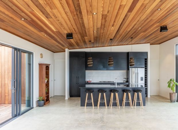 Bright, open-plan kitchen with a large black island and five timber bar stools, dark full-height cabinetry, stainless-steel appliances, and a pale stone-look floor. The room features a striking ceiling clad in warm timber boards with recessed lighting and three sculptural pendant lights. Full-height glazed sliding doors open to an outdoor area, while white walls and abundant daylight create an airy, contemporary atmosphere.