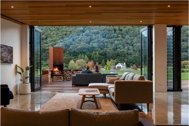 A spacious contemporary living room with floor-to-ceiling retractable glazing opening onto a lush wooded landscape. The room features a warm timber-lined ceiling with recessed spotlights, polished concrete flooring, a large neutral rug, mid-century-inspired sofas and armchairs, and a sculptural indoor fireplace with stacked firewood. The open indoor-outdoor connection and expansive outlook are the dominant features.