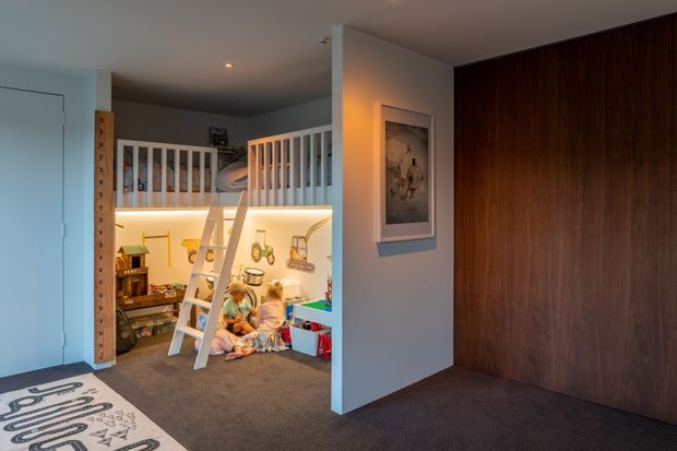A warm, compact children's bedroom or playroom with a raised loft sleeping area, white safety railing, ladder, toys, and a small play space beneath. The room has grey carpet, white walls, recessed lighting, and a large dark timber built-in wall or wardrobe providing contrast.