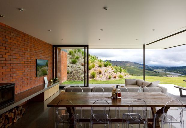 A contemporary open-plan dining and living area with a long timber dining table and transparent acrylic chairs in the foreground, a neutral sectional sofa beyond, and a low media console against an exposed red-brick wall. Full-height glazed sliding doors open the room to a grassy hillside landscape and distant mountains. Recessed ceiling lights, dark flooring, and slim dark window frames create a warm, modern architectural interior.