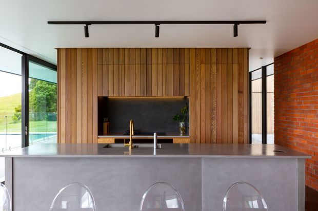 Contemporary open-plan kitchen featuring a large grey island with integrated sink and brass tap, three clear acrylic stools, a warm vertical timber feature wall with built-in dark stone or plaster media niche, black-framed floor-to-ceiling glazing, and an exposed red brick side wall. Track lighting and strong indoor-outdoor views contribute to a refined, modern interior.