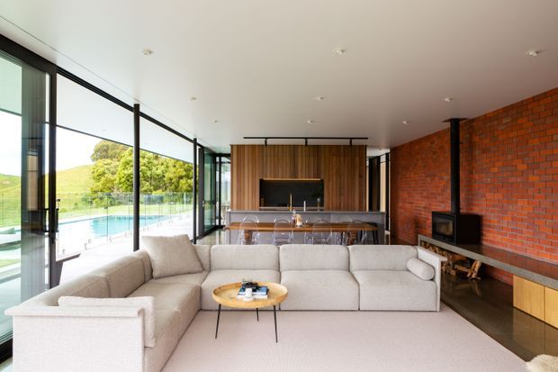 Open-plan contemporary living room with a large cream sectional sofa facing a timber-lined fireplace and integrated media wall. A long kitchen island with stools sits behind the seating area. Full-height black-framed glazing opens to a pool and landscaped outdoors, while an exposed red brick feature wall and dark fireplace add warmth and contrast.