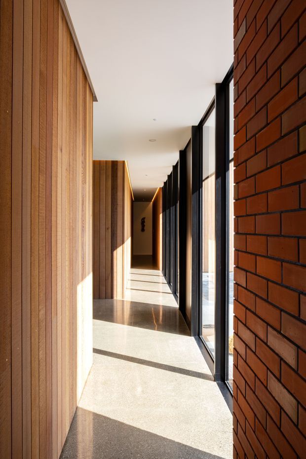 A long, narrow contemporary interior corridor with warm vertical timber wall panelling on one side, exposed red brickwork on the other, and a continuous row of black-framed floor-to-ceiling windows. Polished concrete flooring and strong afternoon sunlight create geometric shadows along the passage, while recessed ceiling lights and concealed linear lighting lead toward a darker end wall.