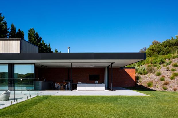 Exterior view of a low, modern flat-roofed house opening onto a manicured lawn and patio. The home features a broad dark roof overhang, extensive floor-to-ceiling glazing, slender dark framing, exposed red brick walls, and an outdoor living area with built-in seating. A swimming pool is partially visible at the left, while a landscaped sloping garden rises along the right side.