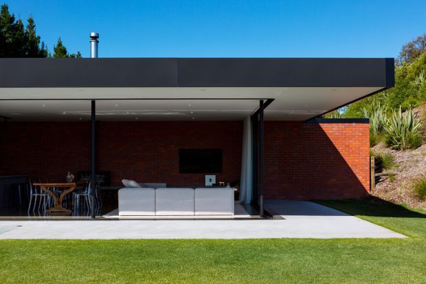 A low-slung modern pavilion or extension with a broad flat roof and deep covered outdoor living area. Dark metal fascia and slender black posts frame a red brick wall, with an outdoor lounge, dining setting, wall-mounted television, and concrete terrace opening onto a neatly maintained lawn. A landscaped sloping garden with established planting appears at the side.