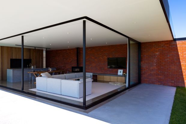 A contemporary open-plan living area viewed from an outdoor paved terrace. Full-height glazed sliding walls and slender dark frames enclose a room with a pale sectional sofa, dining table, compact kitchen, and timber media cabinetry. Exposed red brick walls contrast with the white flat ceiling and concrete terrace, creating a strong indoor-outdoor connection.