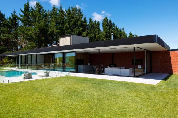 A low, single-storey modern residence set against a backdrop of tall trees, with a broad flat roof, extensive floor-to-ceiling glazing, dark roof and trim, and red brick walls. A covered outdoor living area opens onto a lawn and swimming pool, with glass pool fencing and contemporary outdoor furniture.