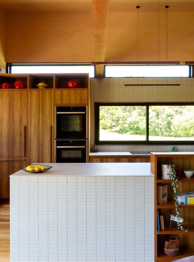Warm, wood-lined contemporary kitchen with a large tiled island, integrated timber cabinetry, double wall ovens, open shelving, and a broad horizontal window overlooking greenery. Fine horizontal wall and island tiles create a strong linear texture, while a timber ceiling and structural post add warmth. Pendant lights hang above the work area, and curated shelving displays ceramics and books.
