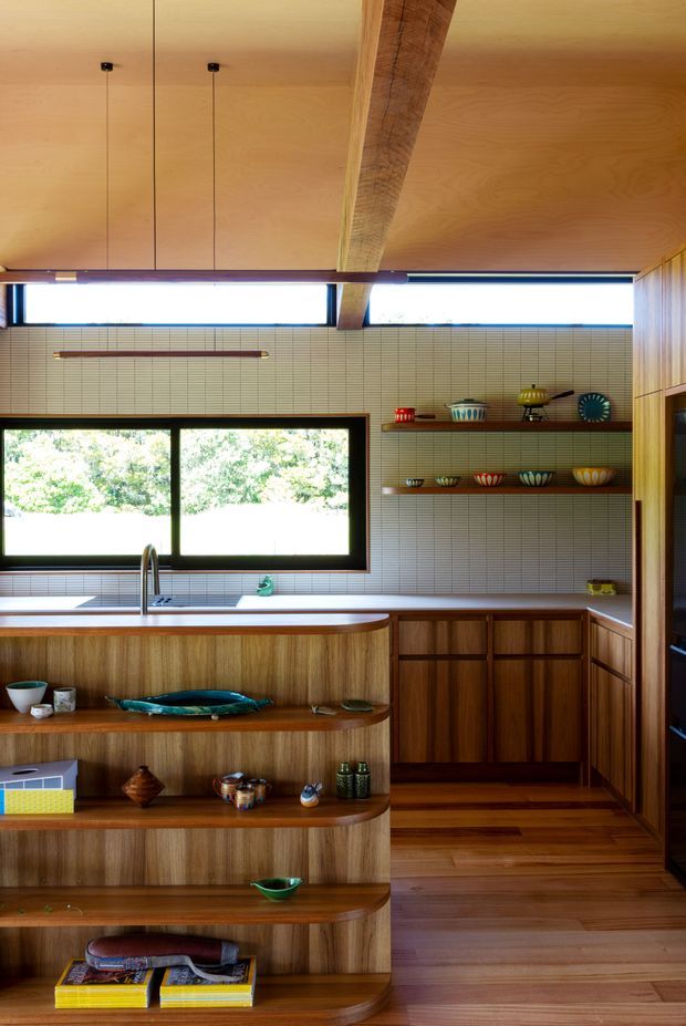 A warm, carefully detailed kitchen with extensive timber cabinetry and shelving, a wood-lined ceiling with an exposed beam, and a tiled backsplash of small pale rectangular tiles. A broad window overlooks greenery, while colourful ceramics and decorative objects line the open shelves and island shelving. The space has a strong mid-century-inspired character with natural materials and abundant daylight.