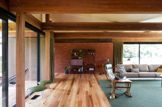 Warm mid-century-inspired living room with exposed timber beams and posts, extensive timber flooring, a red brick feature wall, and large glazed openings. The room includes a sofa, lounge chairs, built-in shelving, and a compact coffee table, with a partially visible adjoining outdoor area beyond the glazing.