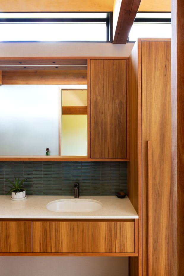 A compact, warm-toned bathroom vanity with extensive timber joinery, a pale solid-surface benchtop, integrated white basin, dark horizontally stacked tile splashback, wall-mounted black tapware, and a large mirror. A high clerestory-style window and exposed dark timber ceiling beam bring natural light into the space.