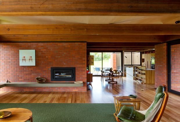 A warm mid-century living room with exposed dark timber beams and ceiling structure, red brick walls, polished timber flooring, and a long built-in brick fireplace. The room opens toward a bright dining area and kitchen, with expansive glazing, timber joinery, and vintage-style furnishings including a green upholstered lounge chair and low coffee table.
